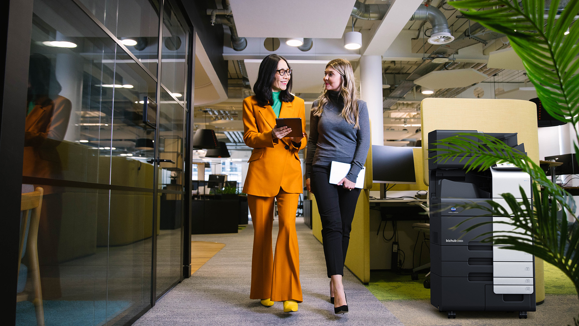 Two women walking through a modern office, discussing work while holding a tablet and notebook, with a Konica Minolta bizhub printer in the background.
