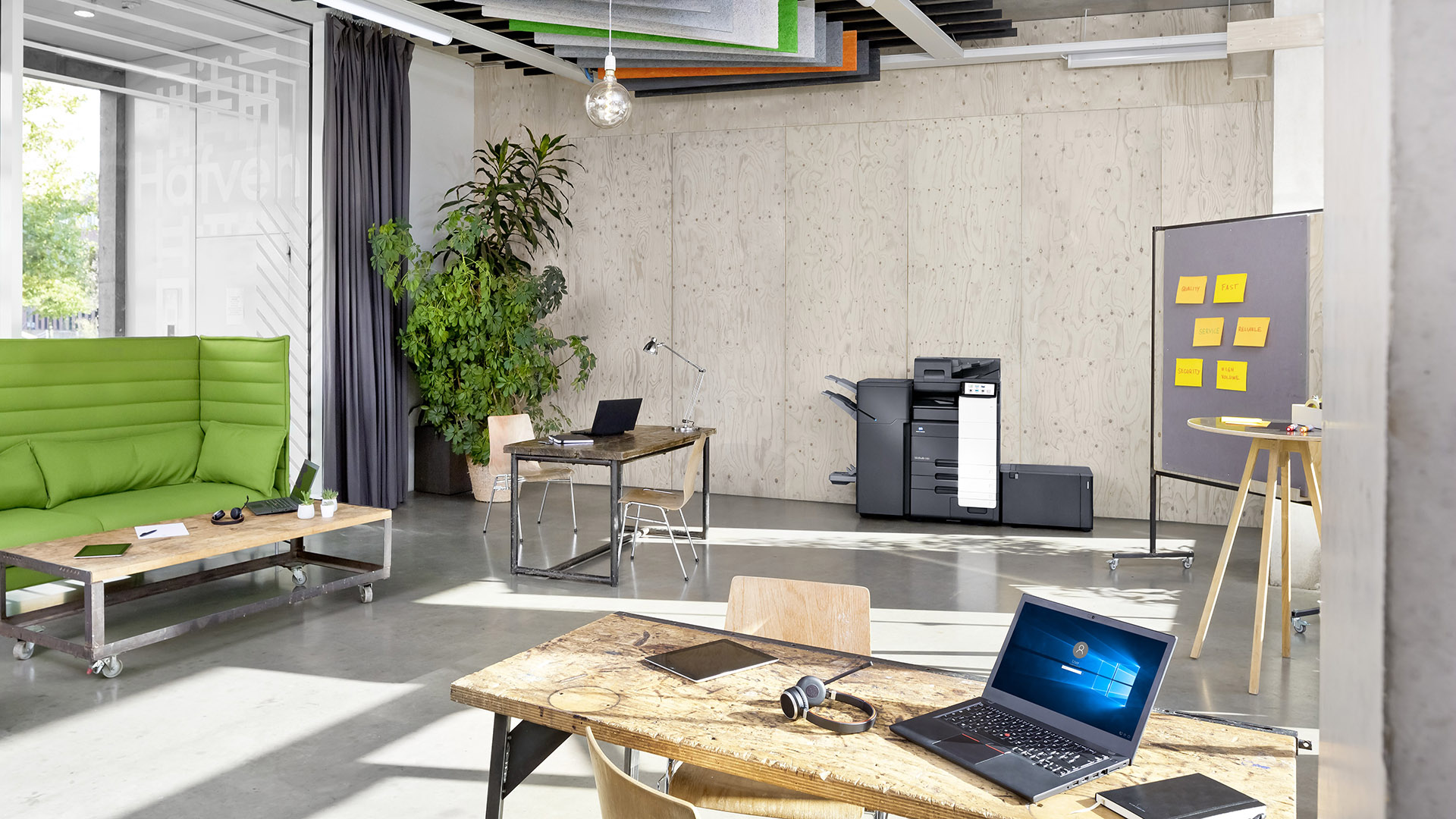 A modern office with natural light and industrial design elements featuring a Konica Minolta bizhub C651i printer against the back wall.