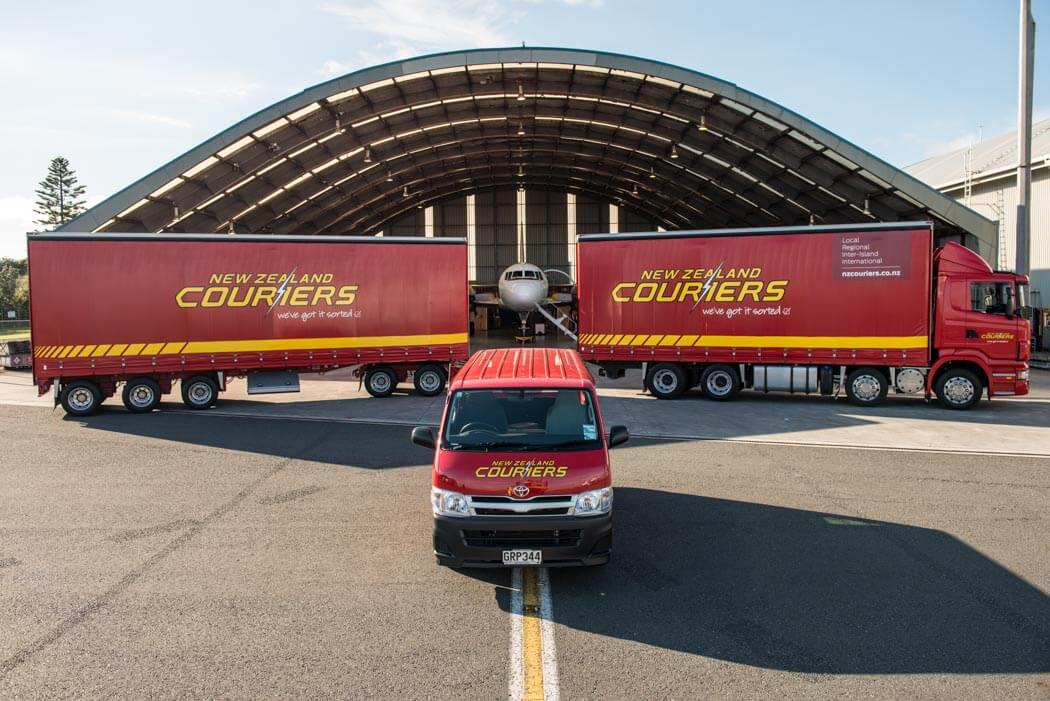 a red New Zealand Couriers van parked in front of a New Zealand Couriers truck, in front of an airplane hanger.