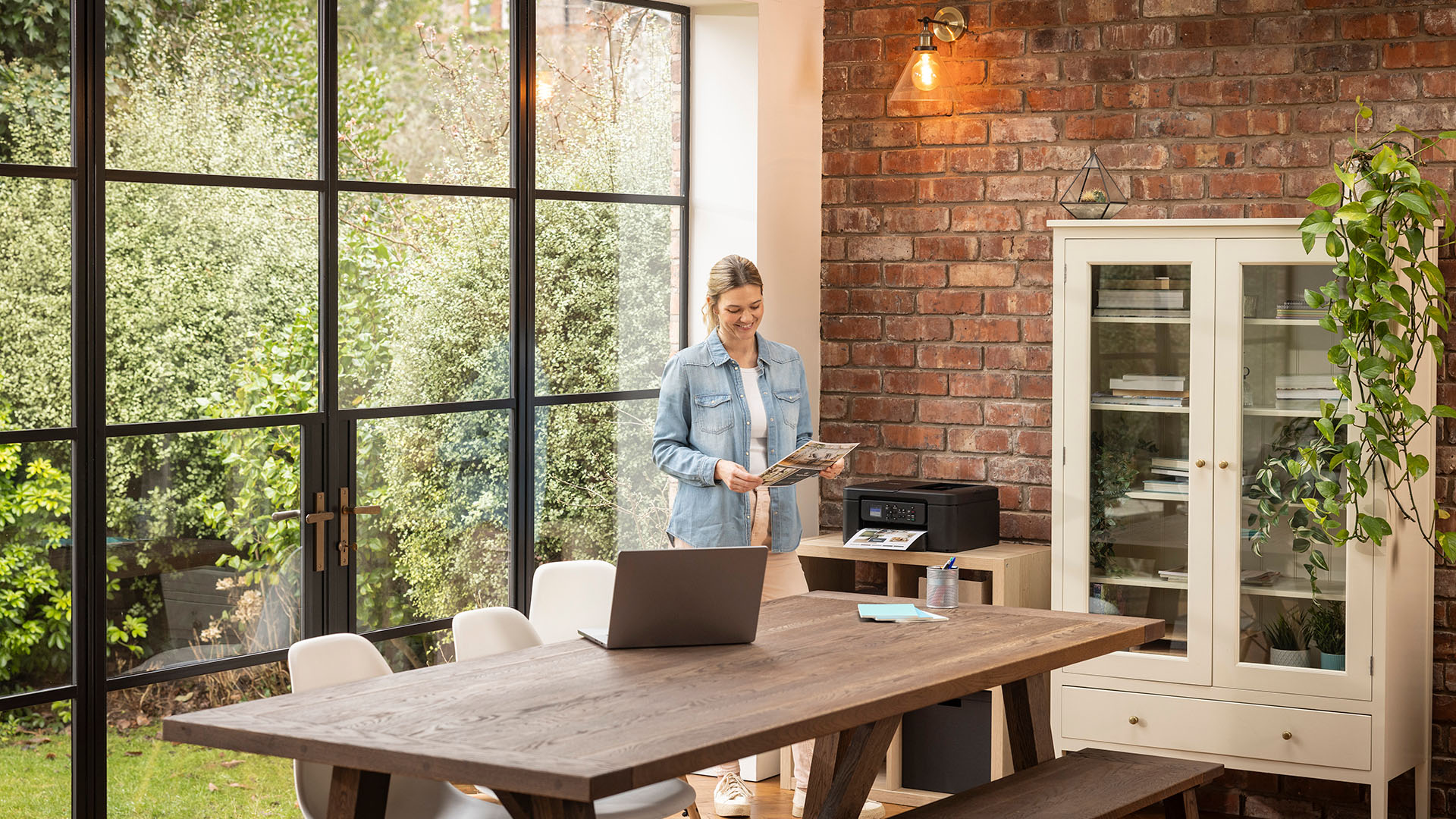 Woman collecting a printed page from a Brother printer in a bright home office with large windows.