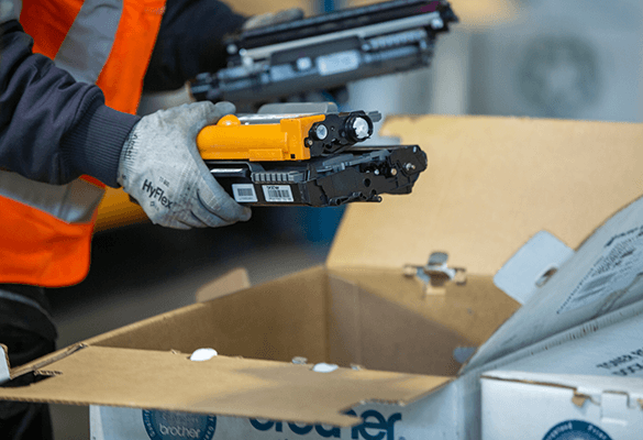 A person wearing a high-visibility orange vest and gray gloves is holding two printer toner cartridges above an open cardboard box labeled 