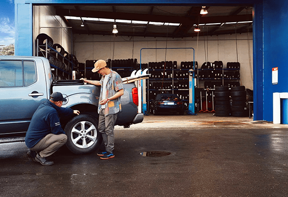 Two men inspecting a gray utility vehicle's tyre at Advantage Tyres, with visible tire racks and equipment inside.