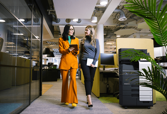 two business women walking through a modern office with a Konica Minolta photocopier against a wall on the right hand side