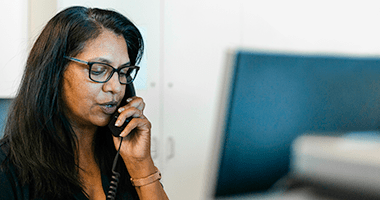 A person with long hair holding a phone to their ear. The person's face is blurred, and they are wearing glasses. The background includes an out-of-focus computer monitor and some office furniture.