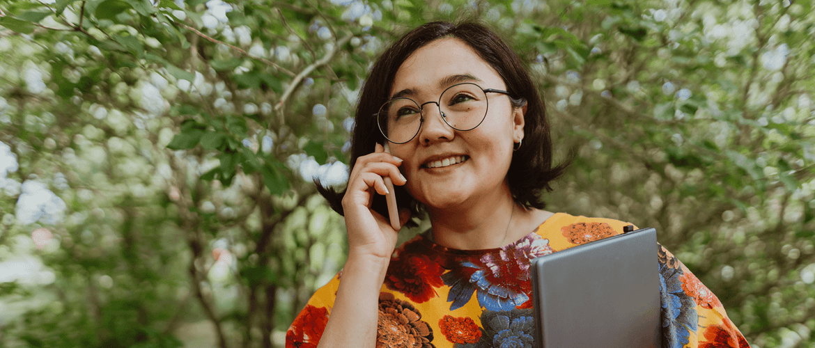 A woman with glasses and short black hair, wearing a colourful floral top, talks on her smartphone while holding a tablet. She is outdoors with green trees in the background, smiling warmly.