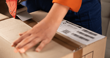 A person sealing a cardboard box with packing tape. The person's hand is pressing down on the tape, and the other hand is holding a tape dispenser. There is a shipping label on the box.
